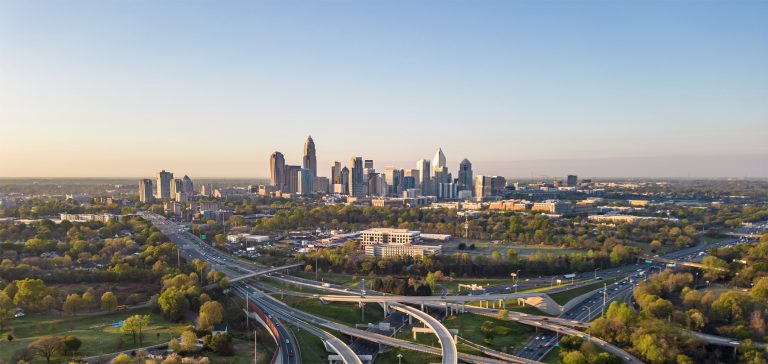 Aerial view of downtown Charlotte, North Carolina, with modern skyscrapers, highways, and surrounding greenery at sunset.